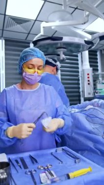 Sarı koruyucu gözlük, şapka, maske ve mavi bornoz takan kadın hemşire cerrahi standındaki aletleri temizliyor. Doktor ameliyatta cerraha yardım ediyor. Dikey video.