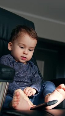 Küçük bebek televizyon kumandasıyla oynuyor. Siyah ofis koltuğunda oturuyor. Evde çıplak ayakla oynayan sakin bir çocuk. Düşük açı. Dikey video