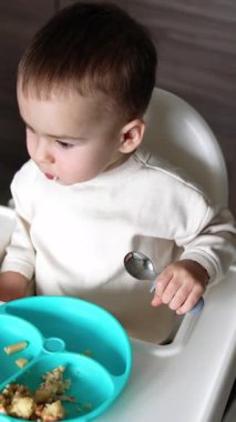 Beyaz gömlekli bebek mavi bir tabağın önünde oturuyor. Elinde kaşık tutan bir çocuk. Anne oğlunu besliyor. Üst Manzara. Dikey video