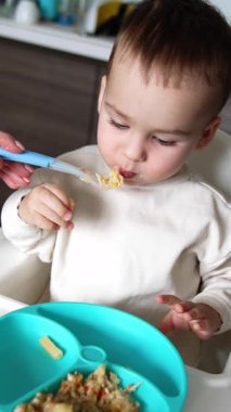 Anne oğlunun ağzına bir kaşık tıkar. Sevimli çocuk masadan bir kaşık alıyor. Kapatın. Mutfak arka planı. Dikey video