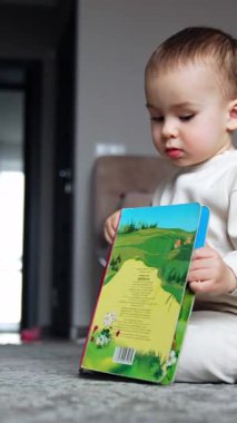 Güzel çocuk renkli bir kitapla oynuyor. Beyaz takım elbiseli sevimli çocuk parlak resimlere bakıyor. Dikey video