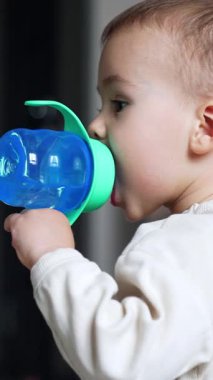 Bebek emzik ile mavi bir şişeden su içiyor. Yan görüş. Bebek merakla yukarı bakıyor. Arkaplan bulanık. Dikey video