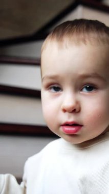 Küçük oğlan çorabını çıkarmak için çok çabalıyor. Merdivenlerde oturan sevimli çocuk kameraya bakıyor. Kapatın. Dikey video