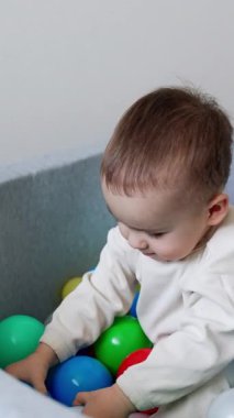Kuru havuzda oynayan şirin beyaz bir bebek. Beyaz takım elbiseli çocuk topu leğenden fırlattı ve oyuncakların üzerine uzandı. Dikey video