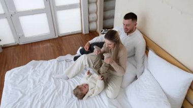 Yatakta oturan sarışın kadın oğlunun bileklerinden tutarak kaldırıyor. Anne bebeğiyle oynuyor. Babam arkada oturuyor. Yüksek açı görünümü.