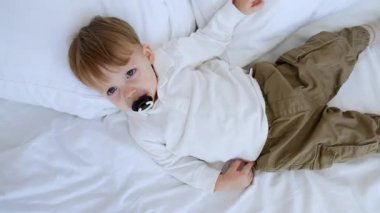 Beyaz gömlekli ve haki pantolonlu beyaz çocuk yatağın üzerinde yatıyor. Bebek emziği emerken yavaşça uykuya dalıyor..