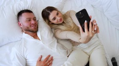 Kafkasyalı çift yatakta birlikte yatıyor. Çekici kadın kendisiyle erkeğinin selfie 'sini çekiyor. Üst görünüm.