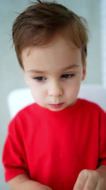 Kırmızı tişörtlü güzel beyaz çocuk yemek çiğniyor. Çocuk portakalı soyuyor, ağzına sokuyor ve neşeyle ellerini kaldırıyor. Dikey video.