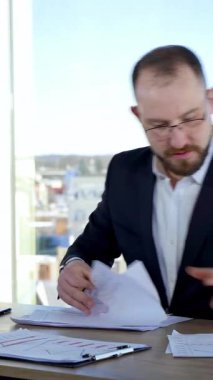 İş yerinde gözlüklü bir iş adamı. Takım elbiseli bir yönetici bir sürü gazeteyle masa başında oturuyor ve pencere arkasındakilere karşı ofiste çalışıyor. Dikey video