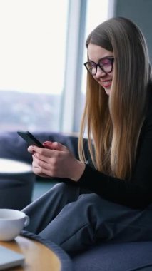Ofisinde telefonu olan güzel bir kadın. Genç iş kadını rahat bir koltukta oturuyor ve pencereye yakın bir telefonla dinleniyor. Laptop ve küçük masada bir fincan. Dikey video