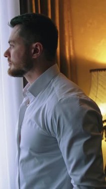 Dairedeki işten sonra yakışıklı bir adam. Karanlık otel odasındaki pencerenin yanındaki genç işadamı. Bir adam otele geldi. Dikey video
