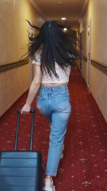 Evinde bavulu olan mutlu bir bayan gezgin. Günlük giysiler içinde genç bir kadın otel koridorunda tramvay çantasıyla yürüyor ve neşeyle zıplıyor. Tatil konsepti. Dikey video