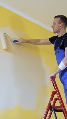 Merdivenden evin duvarlarını boyayan bir işçi. Genç müteahhit erkek duvarları yeni bir açık renge boyuyor. Ev tamiri. Dikey video