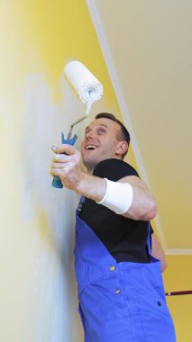 Boya silindirli bir ressam. Dairenin duvarını boyayan mutlu, genç bir adam. İç dekorasyon. Dikey video