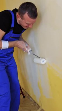 Genç adam bir silindirle beyaz bir duvar boyuyor. İçerisinin tamiri. Erkek dekoratör boş odada bir duvar boyuyor. Tulumlu bir ressam odayı yeniliyor. Dikey video