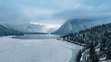 Karlı dağlarla çevrili buzla kaplı göl üzerinde insansız hava aracı görüntüsü. Kayaları kaplayan çam ağaçlarının kış manzarası. Dağların zirveleri gri bulutlarda.