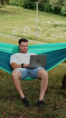 Adam elinde dizüstü bilgisayarla mavi bir hamakta oturuyor. Serbest çalışan adam doğada uzaktan yakından çalışıyor. Dikey video
