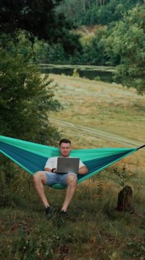 Beyaz adam hamakta sallanarak oturuyor. Freelancer doğada dizüstü bilgisayarında uzaktan kumanda yapıyor. Dikey video