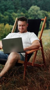 Güzel doğada çalışan mutlu bir erkek serbest yazar. Adam dizüstü bilgisayarıyla katlanan bir sandalyede oturuyor. Dikey video