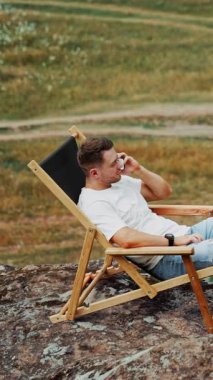 İnsan doğada dinleniyor. Beyaz erkek kayanın üzerinde rahat bir katlanır sandalyede oturup telefonda konuşuyor. Üst Manzara. Dikey video