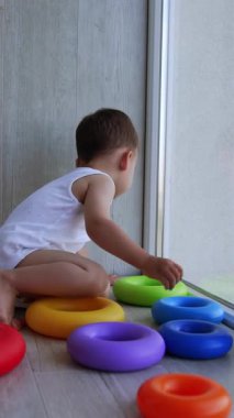 Küçük çocuk oyuncak piramitlerle yere oturmuş etrafa saçılmış. Yeni yürümeye başlayan çocuk kafasıyla cama yaslanmış pencereye bakıyor. Yan görüş. Dikey video
