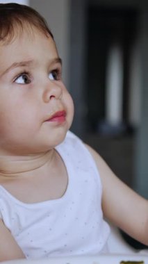 Beyaz elbiseli şirin çocuk yüksek sandalyede oturuyor. Annesi ona kaşıkta yemek veriyor ama inatçı çocuk yemeyi reddediyor. Kapatın. Dikey video