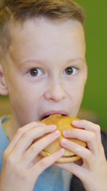 Şehirdeki kafede oturmuş patates kızartması ve hamburger yiyen genç bir çocuk. Fast food kafede bir çocuk. Zararlı beslenme Dikey video