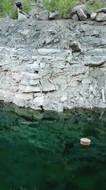 Sırt çantalı, kayalık bir yolda yürüyen genç bir gezginin hava görüntüsü. Gölün kayalık kıyı şeridi ormanla kaplı. Kamp sezonu Dikey video