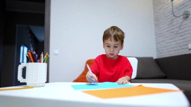 Güzel çocuk masada oturup resim çiziyor. Konsantre çocuk kalem ve renkli kağıt kullanıyor. Düşük açı görünümü.