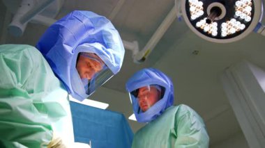 Cüppeli ve koruyucu kasklı cerrahlar travma cerrahisinde işbirliği yaparlar. Operasyon odasında yuvarlak lambanın altında çalışan doktora düşük açılı bakış açısı.