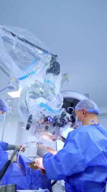 Doktor ameliyat sırasında plastikle kaplı mikroskoba bakıyor. Cerrahın ameliyat yaparken düşük açılı görüntüsü. Hemşire alet cihazını doktora verir. Dikey video.