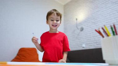 Kırmızı tişörtlü, kalemle çizen mutlu beyaz çocuk. Sanat hobisine odaklanmış bir çocuğun portresi. Düşük açı. Dikey video.