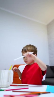 Masada oturan beyaz çocuk kupanın içine kalem koyuyor. Evde çizim yapan küçük bir çocuk. Dikey video.
