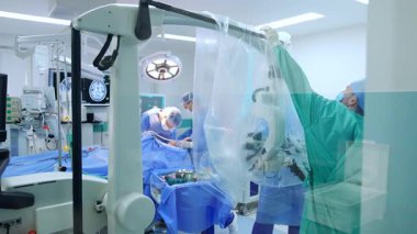 Erkek sıhhiyeciler ameliyat odasındaki mikroskobu sterilite etmek için plastikle kaplıyorlar. Beyin cerrahları arkaplanda ameliyat masasında çalışırlar..