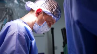 Maskeli ve şapkalı beyaz beyin cerrahı elinde forseps tutuyor. Doktor kesiği dikmek için iplik kullanıyor..