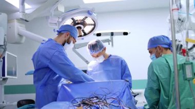 Ameliyat masasında üç üniformalı doktor çalışıyor. Tıbbi cihazlardan gelen birden fazla kablo hastayı koruyan mavi çarşafın üzerinde..