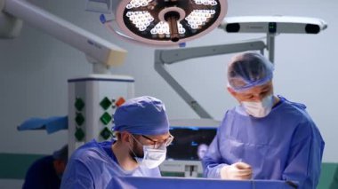 Kafkasyalı doktorlar ameliyat yapıyor. Mavi üniformalı doktorlar, yuvarlak lambayla aydınlatılmış ameliyat yapıyorlar..