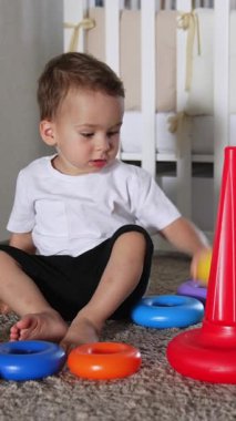 Güzel beyaz çocuk halıda oturup piramitle oynuyor. Bebeğin yanında harika bir siyah kedi yatıyor. Dikey video