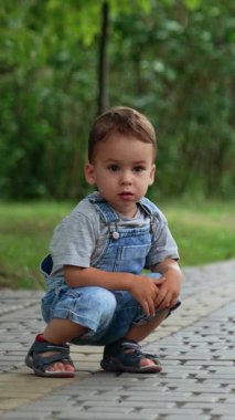 Güzel beyaz çocuk asfalt yolda çömelmiş oturuyor. Çocuk düşünceli bir şekilde ileriye bakıyor ve koşuyor. Dikey video