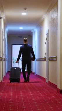 Otel koridorunda bavulla yürüyen işadamlarının arka görüntüsü. Beyaz duvarları, kapıları ve kırmızı halısı olan modern bir iç mekan. Dikey video