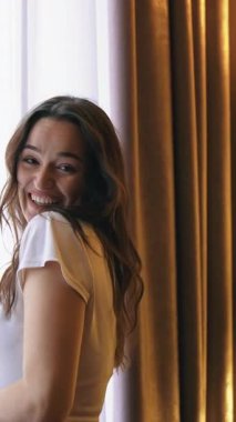 Genç, mutlu, gülümseyen kadın bir otel odasının penceresinden dışarı bakıyor. Modern bir otel odasındaki büyük pencereler güncel olarak tasarlanmış. İyi tatiller. Dikey video