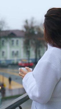 Otel balkonunda elinde bir fincan kahveyle bornozlu güzel bir kızın fotoğrafı. Gelinin sabahı. Genç bir kadın balkonda kahvaltı yapıyor. Dikey video