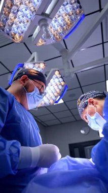 Kadın cerrah ve erkek asistan birlikte çalışıyorlar. Cerrahi odasındaki lambaların altında çalışan doktorlara düşük açılı bakış açısı. Dikey video.