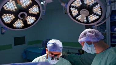 İki profesyonel cerrah mavi çarşafla kaplı hastaya bakıyor. Doktorlar ortopedik ameliyat yaparlar. Parlak yuvarlak lambalar altında sağlık görevlilerine yüksek açı perspektifi.