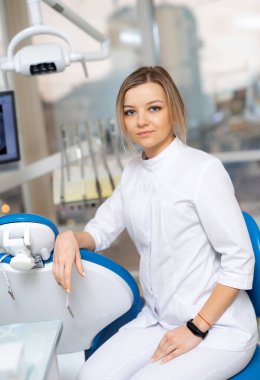 Modern bir klinikte profesyonel bir dişçi. Beyaz önlüklü genç bir dişçi, iyi donanımlı bir dişçi ofisinde güvenle oturur..