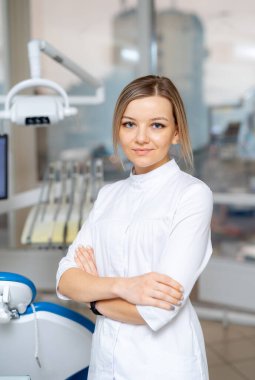 Klinikteki kendine güvenen dişçi. Profesyonel bir diş hekimi, ekipman ve doğal ışıkla dolu modern bir klinikte kollarını kavuşturarak gururla durur..