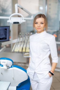 Modern klinikte genç bir dişçi. Bir diş hekimi, gelişmiş ekipmanlar ve sıcak bir atmosferle dolu modern bir klinikte güvenle duruyor..