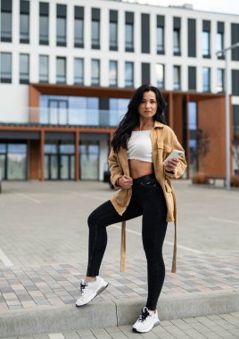 Model, modern bina tarafından poz veriyor. Genç bir kadın şehir ortamında modern mimarinin arka planına karşı şık kıyafetini sergiliyor..