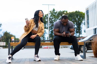 Parkta eğlenen bir çift. Bir adam ve bir kadın parkta bir bankta oturuyorlar, ikisi de telefonlarına odaklanmış bir şekilde birbirlerinin arkadaşlığından keyif alıyorlar..