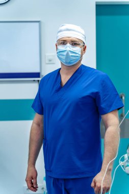 Doktor ameliyata hazırlanıyor. Mavi önlüklü ve maskeli bir tıp uzmanı parlak ışıklı bir ameliyathanede ameliyata hazır bekliyor..